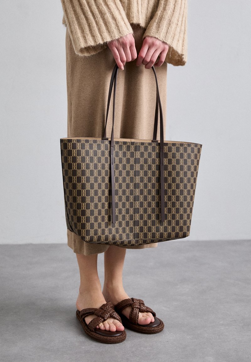 Brown patterned tote bag with black handles, made of durable material. Worn with a beige knitted sweater and strappy brown sandals.