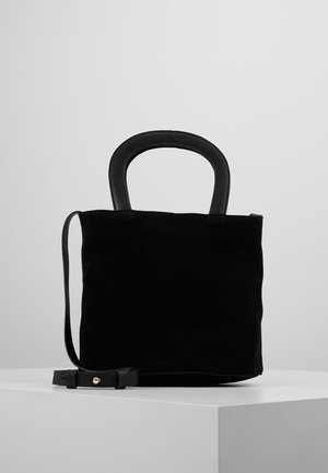 Sac à main en daim noir avec des poignées en cuir arrondies et une fine bandoulière amovible, posé sur un cube blanc devant un fond gris.