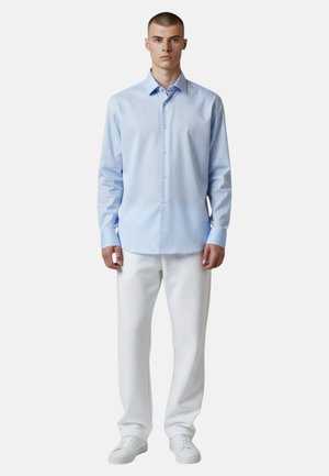 Giovane uomo in piedi che indossa una camicia azzurra con bottoni, pantaloni bianchi e scarpe da ginnastica bianche su uno sfondo bianco semplice.