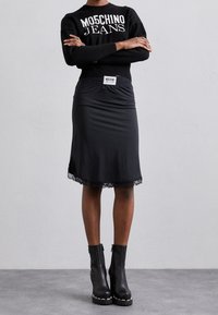 Person klädd i svart Moschino Jeans sweatshirt, svart kjol med spetskant, och svarta platå ankle boots, står med armarna i kors.