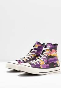Wysokie sneakersy z teksturowanym fioletowym, czarnym i żółtym wzorem kamuflażu, z białymi sznurówkami i gumowymi podeszwami.
