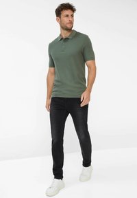 Kaki grøn kortærmet polo skjorte med krave, parret med sorte jeans og hvide sneakers, stående mod en lys baggrund.