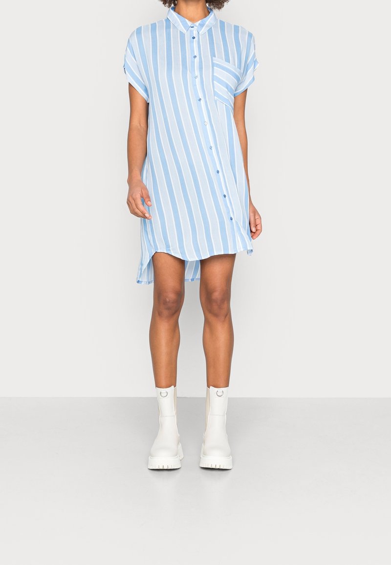 Personne portant une robe chemise à rayures verticales bleu clair et blanc avec des manches courtes et des bottines blanches, debout sur un fond uni.