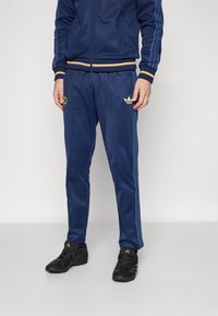 Blauwe tracksuit met gele en rode gestreepte accenten. Bevat een logo op de jack en een klein embleem op de broek. Zwarte sportieve schoenen.