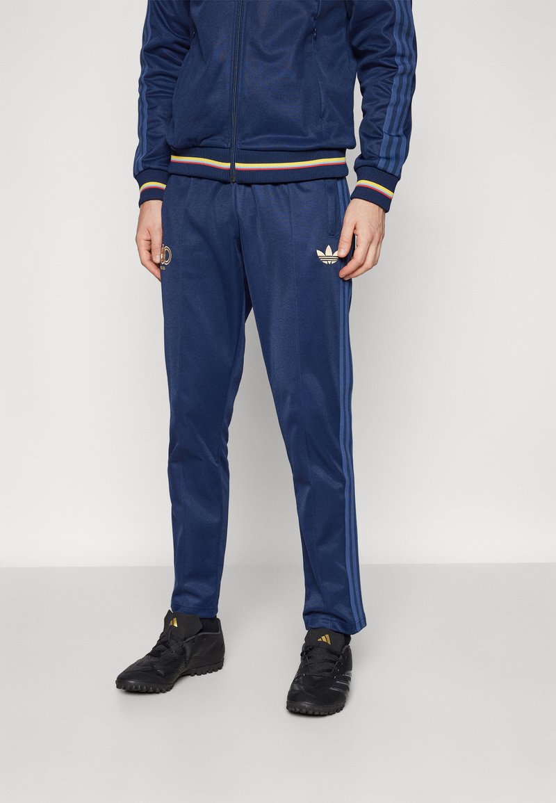 Blauwe tracksuit met gele en rode gestreepte accenten. Bevat een logo op de jack en een klein embleem op de broek. Zwarte sportieve schoenen.