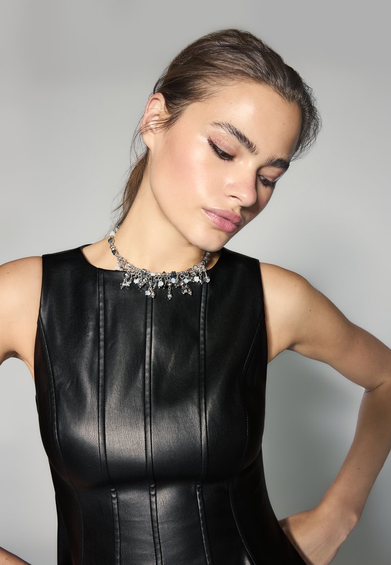 Femme portant une robe en cuir noire ajustée et un collier orné de bijoux pose avec la tête inclinée sur un fond uni gris de studio.