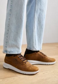 Brązowe skórzane sneakersy z białą podeszwą, z teksturowaną powierzchnią, ciemnymi akcentami i brązowym sznurowadłem, noszone z jasnoniebieskimi dżinsami.