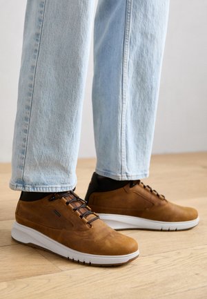 Brązowe skórzane sneakersy z białą podeszwą, z teksturowaną powierzchnią, ciemnymi akcentami i brązowym sznurowadłem, noszone z jasnoniebieskimi dżinsami.