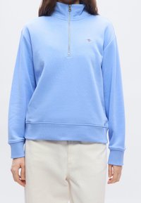 Personne portant un pull à fermeture éclair quart de zip bleu clair et un pantalon écru, montrée des épaules jusqu'aux hanches sur un fond uni.