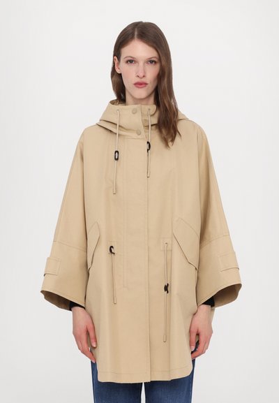 Veste imperméable beige avec une capuche, des cordons de serrage ajustables, des manches larges et des poches latérales. Texture lisse avec des détails de couture minimaux.