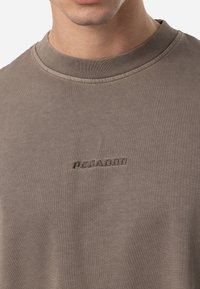 Ruda medvilnės marškinėliai su gurgždančiu kaklu ir iškilusiu "PEGADOR" logotipu krūtinėje, su lygiu, tekstūruotu paviršiumi.