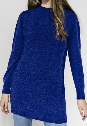 Pull en tricot bleu à manches longues avec un col rond, un ourlet arrière légèrement plus long et une texture douce. Porté avec un jean bleu clair.