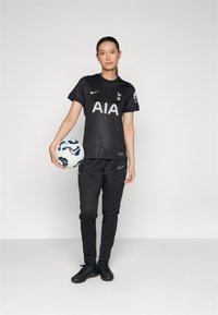 Maillot de football noir avec "AIA" et emblème du club, associé à un pantalon de survêtement noir et des crampons noirs, tenant un ballon de football multicolore.