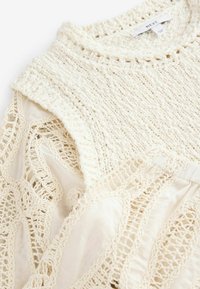 Jasno kremowy sweter wykonany z dzianiny z otwartym splotem, z teksturowanymi nitkami i gładką bazą materiału, prezentujący skomplikowane wzory przypominające koronkę.