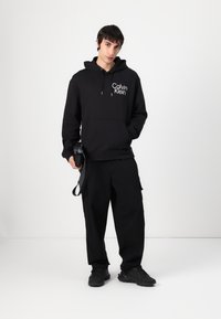Black hoodie with front pocket and Calvin Klein logo, paired with black cargo pants. Model holds a bag with a strap, wears black sneakers.