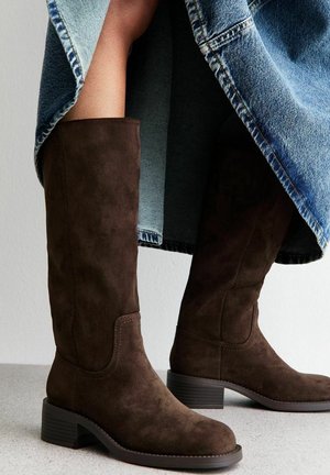 Bottes en daim marron foncé montant jusqu'aux genoux avec des talons bas empilés, portées avec une jupe en jean bleue.