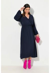 Dunkelblaues langärmeliges Kleid mit V-Ausschnitt und geraffter Taille. Kombiniert mit leuchtend rosa Knöchelstiefeln und einer passenden gepolsterten Clutch.