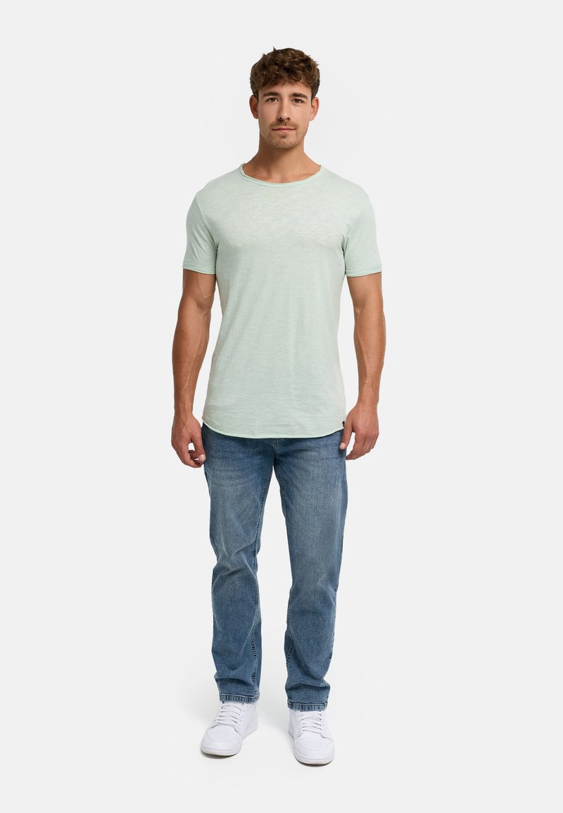 Homme debout face à l’avant, portant une chemise vert clair à manches courtes, un jean bleu et des baskets blanches sur un fond uni.