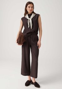 Young woman wearing sleeveless black outfit, white scarf tied at neck, holding brown clutch, standing on plain light background.