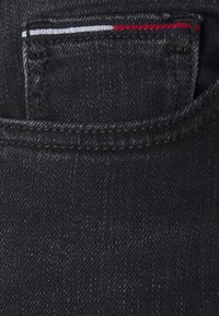 Jeans de mezclilla negra con un pequeño bolsillo frontal y acentos de costura en rojo, blanco y azul. Detalles de textura y costura visibles.