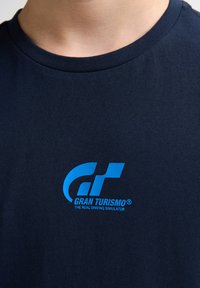 Námořnické bavlněné tričko s kulatým výstřihem, na kterém je světle modré logo "Gran Turismo" a pod ním slogan "Skutečný simulátor jízdy".
