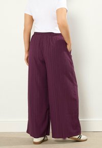 Osoba w luźnych, wysokich fioletowych spodniach z bocznymi kieszeniami i białych sneakersach, stojąca przy białej ścianie.