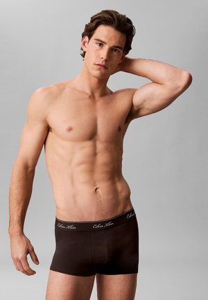 Muskuły mężczyzna pozujący z jedną ręką za głową, ubrany w czarne bokserki Calvin Klein na jednolitym szarym tle.