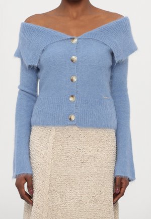 Kvinde iført en lyseblå off-shoulder fluffy cardigan med knapper foran og et cremefarvet tekstureret skørt.