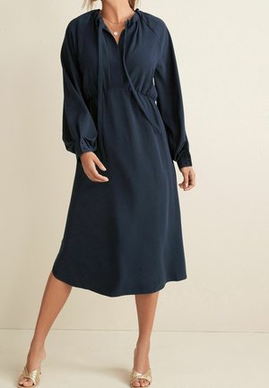 Femme portant une robe midi bleu marine à manches longues, un col à cordon de serrage, et des talons dorés sans brides, debout devant un fond beige.