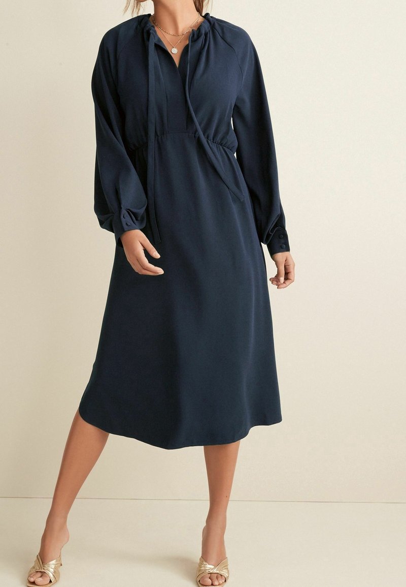 Femme portant une robe midi bleu marine à manches longues, un col à cordon de serrage, et des talons dorés sans brides, debout devant un fond beige.