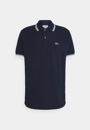 Polo bleu marine à manches courtes avec des doubles rayures blanches sur le col et les poignets, arborant un petit logo crocodile vert sur la poitrine.