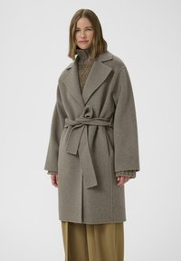 Abrigo de lana gris claro con cintura ceñida por cinturón, grandes solapas y mangas anchas, superpuesto sobre un suéter marrón texturizado y pantalones beige holgados.