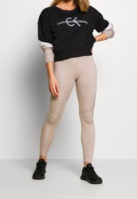 Sweatshirt noir avec logo gris et accents de manches blanches, associé à des leggings beiges avec texte sur le côté. Des baskets de sport noires complètent le look.
