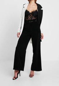 Body de encaje negro con detalles transparentes, combinado con pantalones holgados negros a medida y una chaqueta blanca en contraste. Tacones negros con tiras.