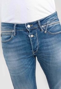 Henkilö, jolla on yllään siniset farkut, joissa on kolme metallinappia, osittain kiinnitettynä, yhdistettynä valkoiseen sisään laitettuun paitaan.