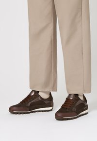 Sneakers in pelle e suede marroni con lacci scuri indossati con pantaloni beige a gamba larga e calzini chiari su uno sfondo bianco.