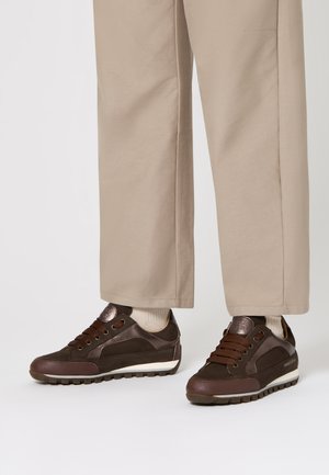 Sneakers in pelle e suede marroni con lacci scuri indossati con pantaloni beige a gamba larga e calzini chiari su uno sfondo bianco.