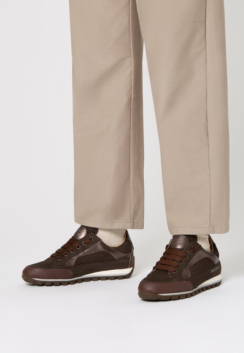 Sneakers in pelle e suede marroni con lacci scuri indossati con pantaloni beige a gamba larga e calzini chiari su uno sfondo bianco.