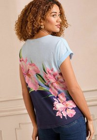 Trumpas rankovėmis pagamintas marškinėlis su mėlyna ir tamsiai mėlyna gradientine spalva, kuriame dideli rožiniai gėlių motyvai ant nugaros. Minkšta medžiaga ir laisvas siluetas.