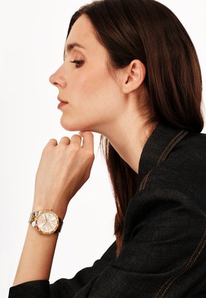 Sølv- og guld armbåndsur med rund skive, romertal og metalarmbånd. Båret på en lys hudfarvet hånd mod en sort jakke.