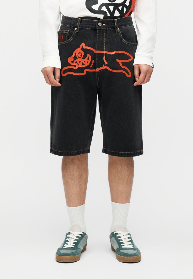 Άτομο που φοράει μαύρα σορτς μέχρι το γόνατο με μεγάλο κόκκινο κινούμενο σχέδιο γάτας μπροστά και τυρκουάζ αθλητικά παπούτσια με λευκά κορδόνια.