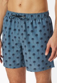 Simshorts i dämpad blått tyg med ett upprepat palmträdsmönster i mörkblått. Elastisk midja med dragsko och sidofickor.