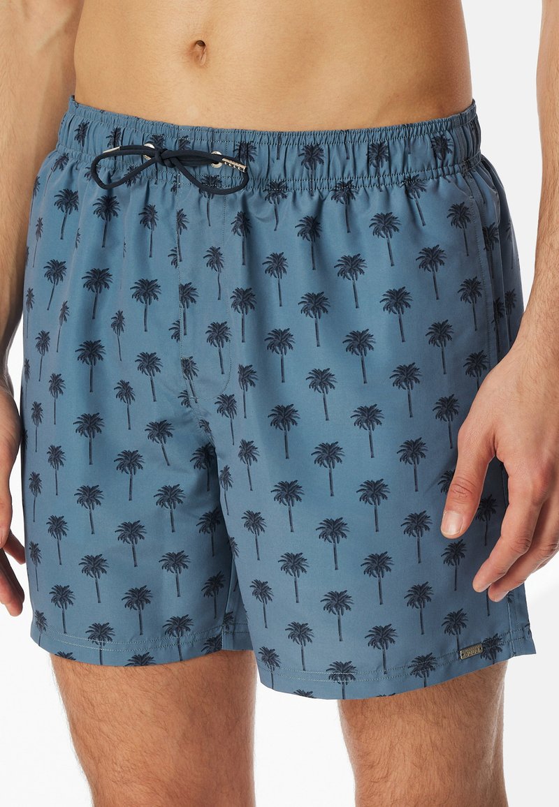 Simshorts i dämpad blått tyg med ett upprepat palmträdsmönster i mörkblått. Elastisk midja med dragsko och sidofickor.