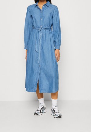 Kvinde iført en langærmet denimkjole med knapper og bånd om taljen, hvide sokker og sporty sorte og hvide sneakers, stående foran en ensfarvet baggrund.