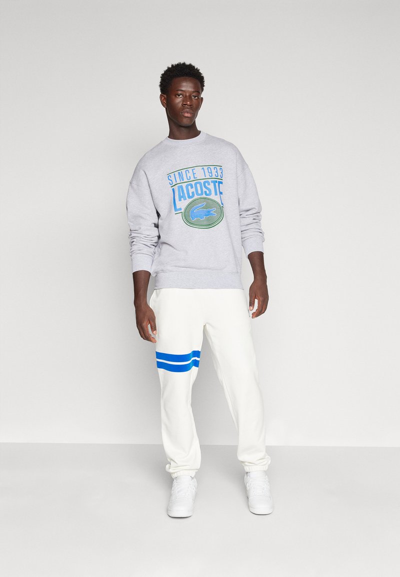 Felpa grigia con logo Lacoste blu e verde, abbinata a pantaloni sportivi bianchi con strisce blu sul lato. Sneakers bianche.