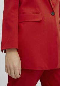 Blazer rosso con colletto a punte, chiusura a un bottone e tasche con pattina. Il tessuto appare liscio e texturizzato con una silhouette aderente.
