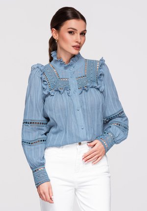 Femme en blouse bleu clair à manches longues avec des détails en dentelle et volants, associée à un pantalon blanc taille haute, sur un fond uni.
