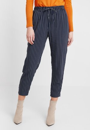 Pantalons à rayures navy avec une taille élastique et un cordon de serrage. Caractéristiques des jambes fuselées et un tissu léger. Portés avec des bottines beiges.