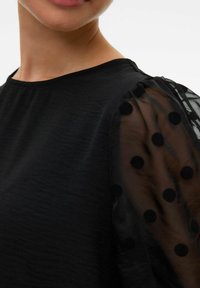 Mustrete top ümmarguse kaelusega ja tekstuurse rinnaosaga. Läbipaistvad mustad puhv-sleeved varrukad, millel on ringide muster. Satinjat sära.