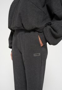 Donkergrijze, getextureerde tracksuit met een ritsjack en broek met elastische taille. Subtiele glinsteraccent en merkpatch op het been.